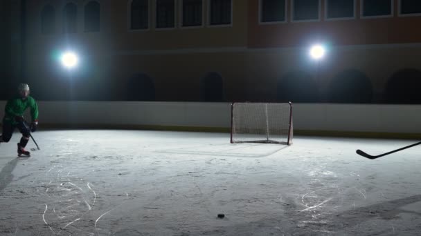 Dos hombres en uniformes y cascos con palos de hockey patinan en la arena de hielo, golpeando el disco y chocan entre sí. Los jugadores profesionales de hockey entrenan en una pista oscura con focos. Movimiento lento. — Vídeos de Stock