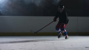 Hokey oyuncusu spot ışıkları ve dumanla karanlık bir buz arenasında kayıyor, hokey sopasıyla buza çarpıyor ve kameraya buz tozu atıyor. Ağır çekim. Kapat..
