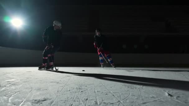 Two sportsmen hockey players who are sliding on ice arena and dribbles, hitting puck with stick. Young guys in uniforms, helmets and with sticks are skating in dark rink with spotlights. Slow motion. — Stock Video