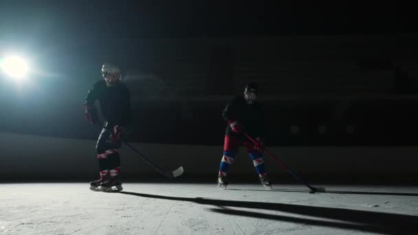 Two sportsmen hockey players who are sliding on ice arena and dribbles, hitting puck with stick. Young guys in uniforms, helmets and with sticks are skating in dark rink with spotlights. Slow motion. — Stock Video