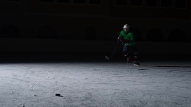 Vorwärts in grüner Uniform und mit Helm und Stock in der Hand rutscht er aufs Eis und trifft den Puck. Ein Sportler läuft auf dem Eis einer dunklen Arena mit Gegenlicht. Zeitlupe. — Stockvideo