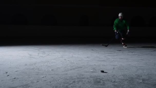 Avanti in uniforme verde con bastone in mani fa scorrere su ghiaccio e colpisce il disco. Uomo scivolare sulla pista e tagliare il ghiaccio in polvere. Un atleta pattina su ghiaccio di arena scura con retroilluminazione. Rallentatore — Video Stock