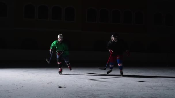 Zwei Männer in Uniformen und Helmen mit Hockeyschlägern laufen über die Eisarena. Ein Spieler schiebt den anderen, dribbelt und trifft den Puck. Hockeyprofis trainieren auf einer dunklen Eisbahn mit Scheinwerfern. Zeitlupe — Stockvideo