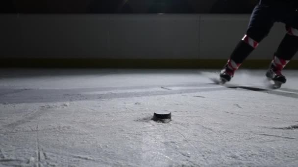Câmera seguindo os pés do jogador de hóquei em patinadores deslizando no gelo. Feche o disco batendo com pau e marcando gol. Esportista patinando na arena gelada escura com holofotes. Movimento lento. — Vídeo de Stock
