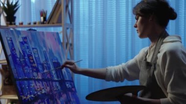 Yetenekli ressam yağlı boya tablolarda çalışıyor, fırça kullanıyor, evleri olan modern şehirler çiziyor. Tuvalin sehpanın üzerinde durduğu karanlık yaratıcı stüdyo. Sanat okulunda ders. Ağır çekim. Kapat..