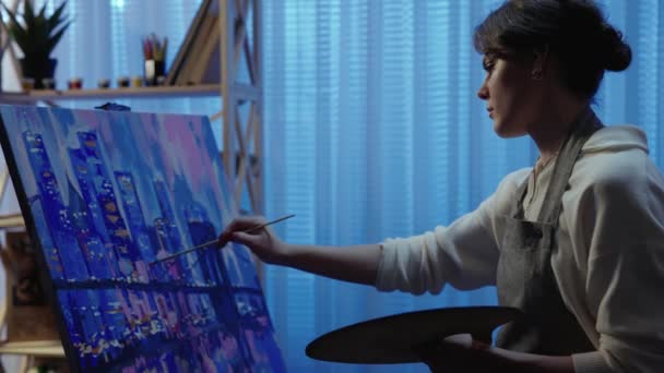 Talented artist works on oil painting, using a brush, she draws modern city with houses. Dark creative studio where the canvas stands on an easel. Lesson in art school. Slow motion. Close up. — Stock Video