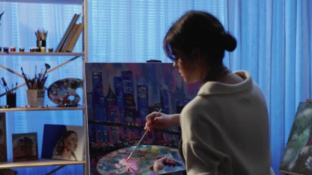 View from back of an artist painting picture on canvas in creative workshop. Young woman creates painting using brush and oil paints on palette. City with buildings close up. Slow motion. — Stock Video