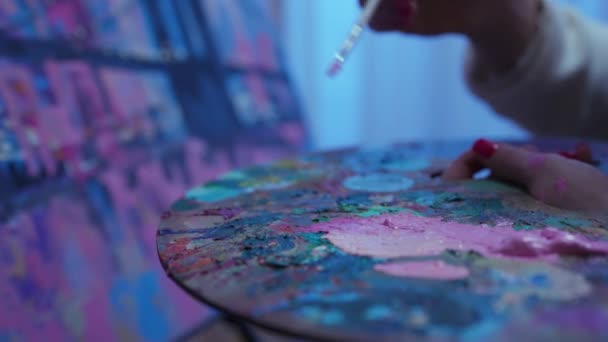 Εστίαση στην παλέτα με ελαιογραφίες. Καλλιτέχνης ζωγραφίζει σε καμβά στο δημιουργικό εργαστήριο. Γυναίκα βουτάει πινέλο σε φωτεινά ακρυλικά χρώματα στην παλέτα και εφαρμόζει πινελιές στη ζωγραφική. Αργή κίνηση. Κλείσε.. — Αρχείο Βίντεο