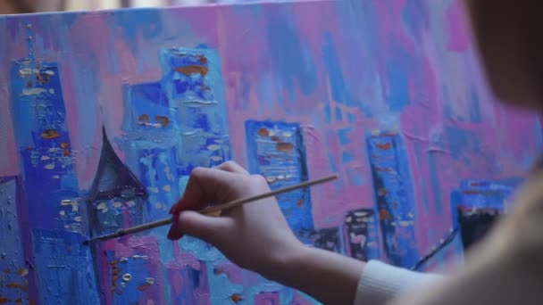 Udsigt fra bagsiden af et kunstnermaleri billede på lærred i kreativt værksted. Ung kvinde skaber maleri ved hjælp af børste og olie maling på palet. By med bygninger tæt på. Langsom bevægelse. – Stock-video