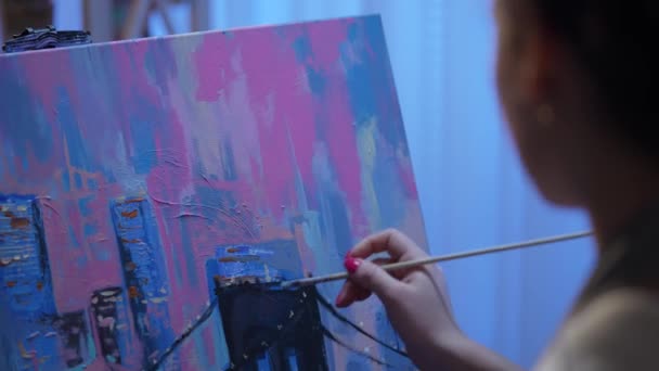 Bekijk vanaf de achterkant van een schilderij van een kunstenaar op doek in een creatieve werkplaats. Jonge vrouw maakt schilderen met penseel en olieverf op het palet. Stad met gebouwen dichtbij. Langzame beweging. — Stockvideo