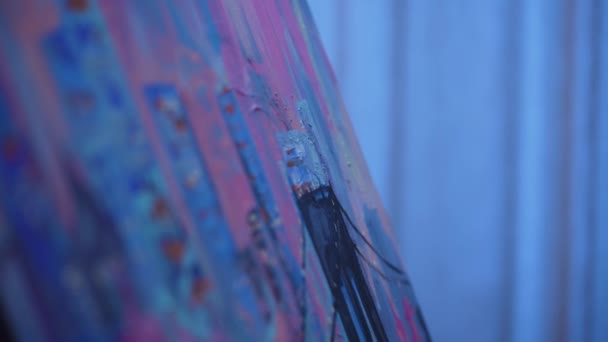 Schilder schilderij met olieverf op doek met behulp van tbrush. Kunstenaar past slagen toe op het beeld met blauwe verf, tekent stad met huizen. Artistieke ambacht in creatieve werkplaats. Langzame beweging. Sluiten.. — Stockvideo