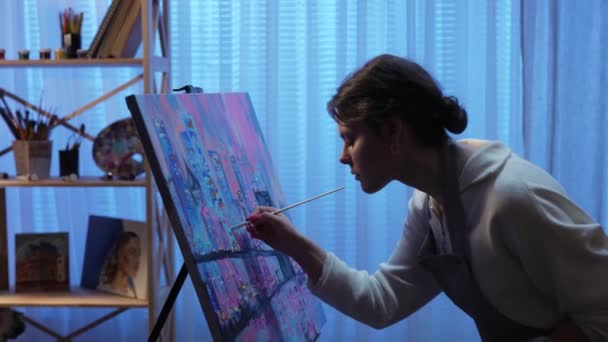 Kunstenaar schilderij op doek in creatieve werkplaats. Jonge vrouw maakt schilderen met penseel en olieverf op het palet. Stad met hoge gebouwen en brug in blauwe tinten close-up. Langzame beweging — Stockvideo