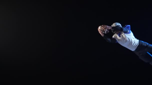 Американский футболист прыгает с мячом в руках и падает на землю. Спортсмен в шлеме, форме и прокладках учится бросать и ловить мяч в темноте на черном фоне арены. Медленное движение. — стоковое видео