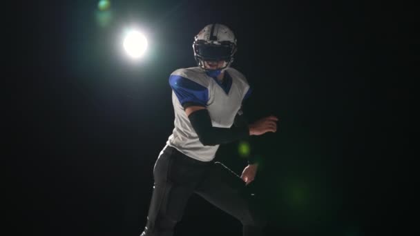 Jogador de futebol americano vestindo uniforme, almofadas e capacete pega passar e corre em ataque contra fundo preto com holofotes. Os jogadores correm atacando para marcar pontos de touchdown. Movimento lento. — Vídeo de Stock