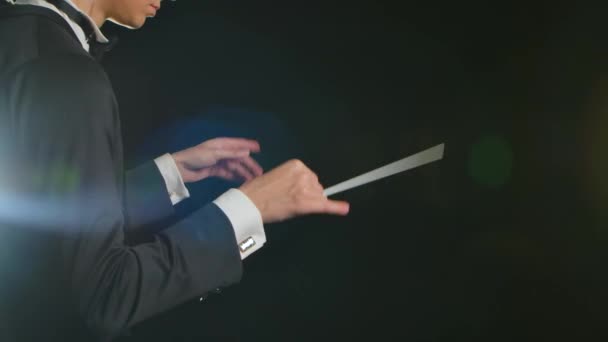 Le chef d'orchestre symphonique portant le costume dirige les musiciens avec le mouvement du bâton sur le fond noir avec les lumières. Direction, mise en scène d'une performance musicale avec des gestes visibles. Gros plan. — Video