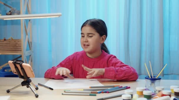 Masada oturan küçük bir kız keçeli kalemlerle resim çiziyor ve telefonda görüntülü konuşuyor. Genç kız yaratıcılık ve çevrimiçi çizim kurslarına katılıyor. Kapatın. Yavaş çekim hazır, 4K 59.94fps 'de. — Stok video