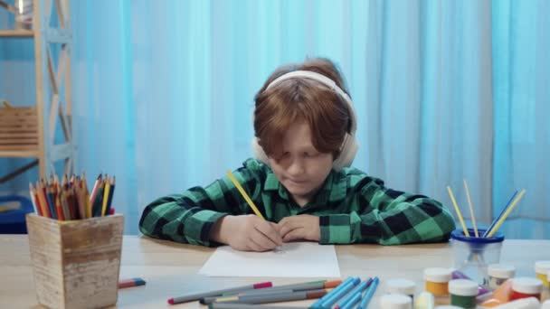 小さな男の子は、部屋のテーブルに座って、アルバムに鉛筆で描画し、大きな白いヘッドフォンで音楽を聴く。10代の少年は創造性に従事している。閉めろ。スローモーション準備完了、 59.94fpsで4K — ストック動画