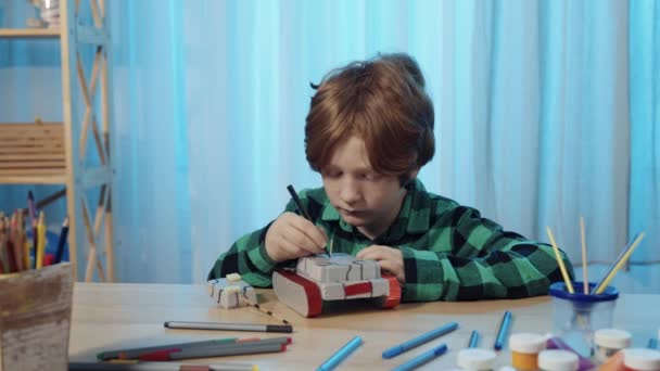 Pequeño colegial sentado en la mesa en la habitación y pinta tanque de juguete. Un adolescente se dedica a la creatividad, dibuja un cuadro, afición, desarrollo, ocio. De cerca. Cámara lenta lista, 4K a 59.94fps. — Vídeo de stock