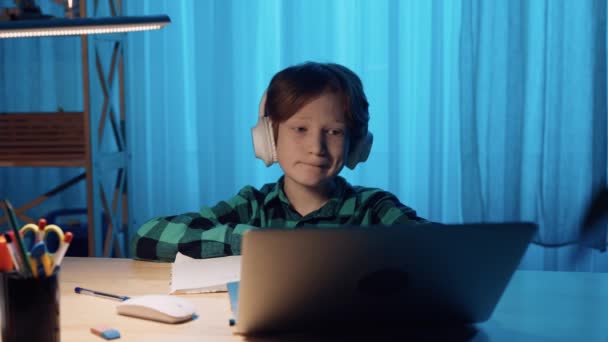 Liten pojke studerar online med hjälp av videosamtal på bärbar dator. Pojken lyssnar på lärarinformation i hörlurar och skriver ner den i anteckningsboken. Närbild. Slow motion klar, 4K vid 59.94fps. — Stockvideo
