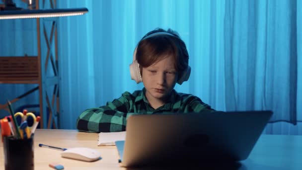 Liten pojke studerar online med hjälp av videosamtal på bärbar dator. Pojken lyssnar på lärarinformation i hörlurar och skriver ner den i anteckningsboken. Närbild. Slow motion klar, 4K vid 59.94fps. — Stockvideo
