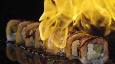 Somon ve unagi soslu Philadelphia suşi rulosu ateşte kavrulmuş. Yangın, siyah arkaplandaki suşi setini kapatır. Somon, pirinç, avokado ve peynirli suşi ızgarası. Yavaş çekim.