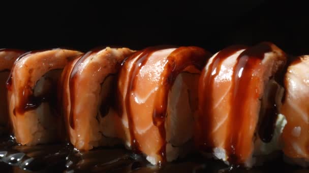 Sushi Filadélfia rolo com molho unagi, girando sobre fundo preto de perto. Rolo de sushi com salmão, arroz, abacate e queijo. Comida tradicional japonesa com frutos do mar e peixe. Movimento lento. — Vídeo de Stock