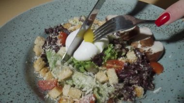 Kadınların elleri, Sezar salatasında haşlanmış yumurtayı çatal ve bıçakla keser. Hindi, domates, marul, kızarmış ekmek, peynir ve sosla kaplayın. Yumurta sarısı sebzelerin üzerine akar. Yavaş çekim.