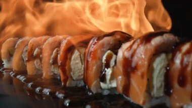 Somon ve unagi soslu Philadelphia suşi rulosu ateşte kavrulmuş. Yangın, siyah arkaplandaki suşi setini kapatır. Somon, pirinç, avokado ve peynirli suşi ızgarası. Yavaş çekim.