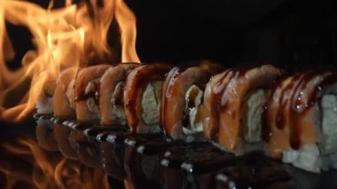 Somon ve unagi soslu Philadelphia suşi rulosu ateşte kavrulmuş. Yangın, siyah arkaplandaki suşi setini kapatır. Somon, pirinç, avokado ve peynirli suşi ızgarası. Yavaş çekim.