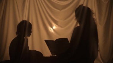 Masal kitabı okuyan kadın ve çocuğun siluetleri. Anne ve küçük oğlu evde perdenin arkasında birlikte vakit geçiriyorlar. Gölgelerin kukla tiyatrosu, gölge ve ışığın oyunu. Kapat..