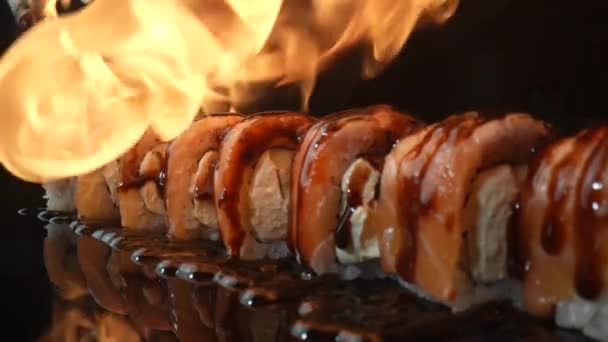 Philadelphia-Sushi-Rolle mit Lachs und auf Feuer gebratener Unagi-Sauce. Feuer umhüllt ein Sushi-Set auf schwarzem Hintergrund in Großaufnahme. Sushi-Rollbraten mit Lachs, Reis, Avocado und Käse. Zeitlupe. — Stockvideo