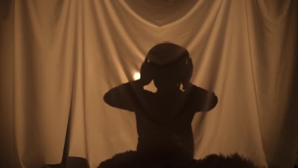 Silhouette di bambino godendo la musica con grandi cuffie. Bambino di età primaria trascorrere il tempo libero, seduto dietro la tenda a casa. Teatro delle marionette delle ombre, gioco delle ombre e della luce. Da vicino.. — Video Stock