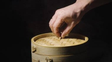 Bir adamın eli bambu buharlı tencerenin kapağını açar ve Çin mantısı wontonunu gösterir, siyah arka planda yakın plan. Çin lokantası mutfağı. Asya buharlı yemeği Dim Sum, doldurulmuş börek. Yavaş çekim.
