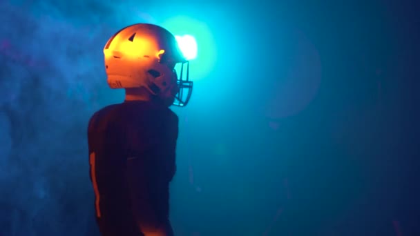 Vista posteriore di un atleta in casco e uniforme che si prepara a giocare a football americano. L'uomo si allena in un'arena buia con luci blu e fumo. Uomo sicuro e aggressivo in uniforme. Da vicino.. — Video Stock