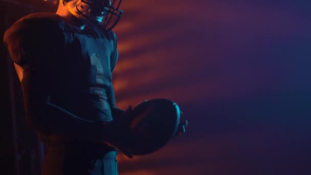 Porträtt av beslutsam professionell amerikansk fotbollsspelare som håller bollen och redo för spel. Mannen poserar i mörka arenan med ljus och rök. Självsäker man i uniform och hjälm. Närbild. — Stockvideo