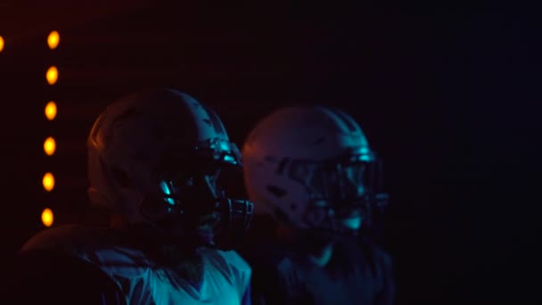 Två beslutsamma idrottare i uniformer och hjälmar redo att spela amerikansk fotboll. Självsäkra och aggressiva spelare utmana motståndare när de står i mörka arenan med ljus. Närbild. — Stockvideo