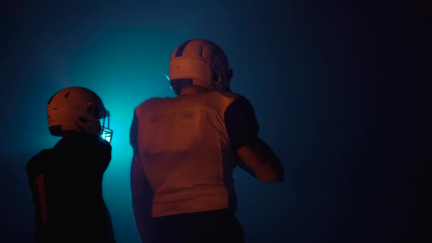 Blick von hinten auf zwei entschlossene Athleten in Uniformen und Helmen, die bereit sind, American Football zu spielen. Selbstbewusste und aggressive Spieler fordern Gegner heraus, während sie in einer dunklen Arena mit Lichtern stehen. Nahaufnahme. — Stockvideo
