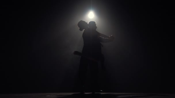 El hombre y la mujer realizan danza latina en la oscuridad con luz y humo. Siluetas de pareja de bailarines bailando apasionadamente tango sobre fondo oscuro. Cámara lenta lista, 4K a 59.94fps. — Vídeo de stock