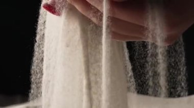 Kadınların elleri avuçlarında kum topluyor ve parmaklarından kum taneleri dökülüyor. Genç bir kadının elleri izole edilmiş siyah stüdyo arka planında beyaz kuru kumla oynuyor. Kapatın. Yavaş çekim