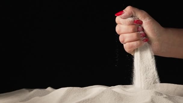 Mão feminina espremendo e derramando punhado de areia seca branca em fundo estúdio preto isolado. Partículas de areia a peneirar os dedos. Close-up de grãos de quartzo mineral natural puro. Movimento lento — Vídeo de Stock