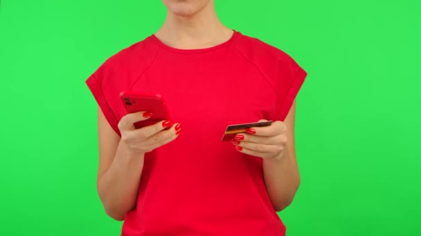 Mujer en camiseta roja hace una compra en línea con una tarjeta de crédito y un teléfono inteligente con espacio de trabajo pantalla simulada sobre fondo verde. Área de publicidad. Lugar de la plantilla. De cerca.. — Vídeo de stock