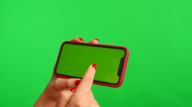 Bir kadın cep telefonunu elinde tutuyor ve ekranı taklit ediyor. Elinde yatay pozisyonda yeşil arka plan var. Kadın eli dokunuyor, tıkırdıyor, tıkırdıyor, tıkırdıyor. Şablon yeri. Kapat.