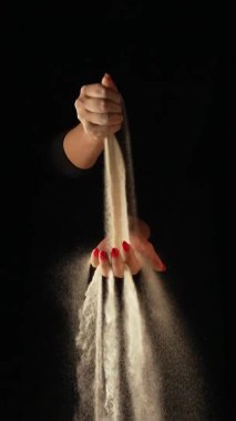 Kuru kum akıntısı bir kadının avucuna dökülüyor ve parmaklarından siyah arka plana dökülüyor. Küçük kum taneleri yavaş çekimde seçilir ve farklı yönlere dağılır. Kapat..
