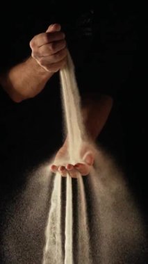 Kuru kum deresi insan avucunun içine dökülüyor ve parmaklarından siyah arka plana dökülüyor. Küçük kum taneleri yavaş çekimde seçilir ve farklı yönlere dağılır. Kapat..