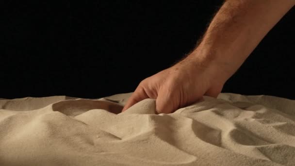 Main masculine serrant et renversant une poignée de sable sec sur fond de studio noir. Particules de sable tamisées à travers les doigts. Gros plan de grains de quartz minéral naturel pur à la main masculine. Mouvement lent. — Video