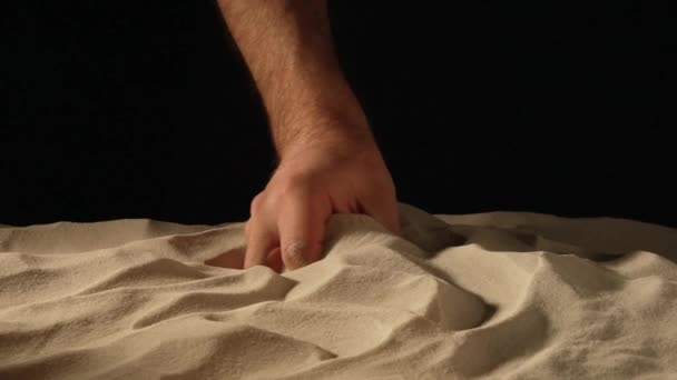 Мужская рука сжимает и проливает горсть сухого песка на черном фоне студии. Частицы песка просеиваются сквозь пальцы. Крупный план зерен чистого природного минерального кварца в мужской руке. Медленное движение. — стоковое видео