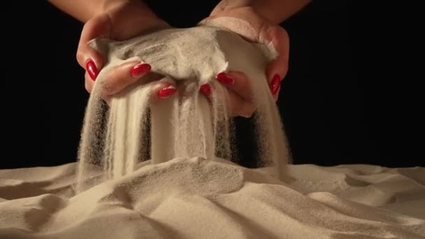 Γυναικεία χέρια μαζεύουν άμμο στις παλάμες τους και χύνουν κόκκους άμμου μέσα από τα δάχτυλά τους. Τα χέρια της νεαρής γυναίκας παίζουν με λευκή ξηρή άμμο σε απομονωμένο μαύρο φόντο στούντιο. Κλείσε. Αργή κίνηση — Αρχείο Βίντεο