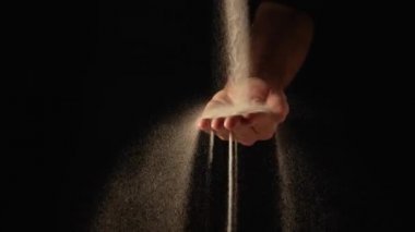 Kuru kum deresi insan avucunun içine dökülüyor ve parmaklarından siyah arka plana dökülüyor. Küçük kum taneleri yavaş çekimde seçilir ve farklı yönlere dağılır. Kapat..