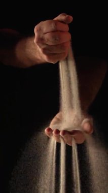 Kuru kum deresi insan avucunun içine dökülüyor ve parmaklarından siyah arka plana dökülüyor. Küçük kum taneleri yavaş çekimde seçilir ve farklı yönlere dağılır. Kapat..