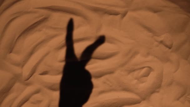 La sombra de una mano de mujer en la arena de la playa mostrando dos dedos gesto de victoria. Cierra la silueta de una mano femenina. Vacaciones de verano junto al mar, vacaciones disfrutando de la playa. Movimiento lento. — Vídeo de stock